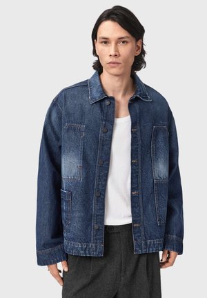 Man met middellang donker haar, gekleed in een donkerblauw spijkerjack met knopen over een wit overhemd en donkergrijze broek tegen een effen achtergrond.