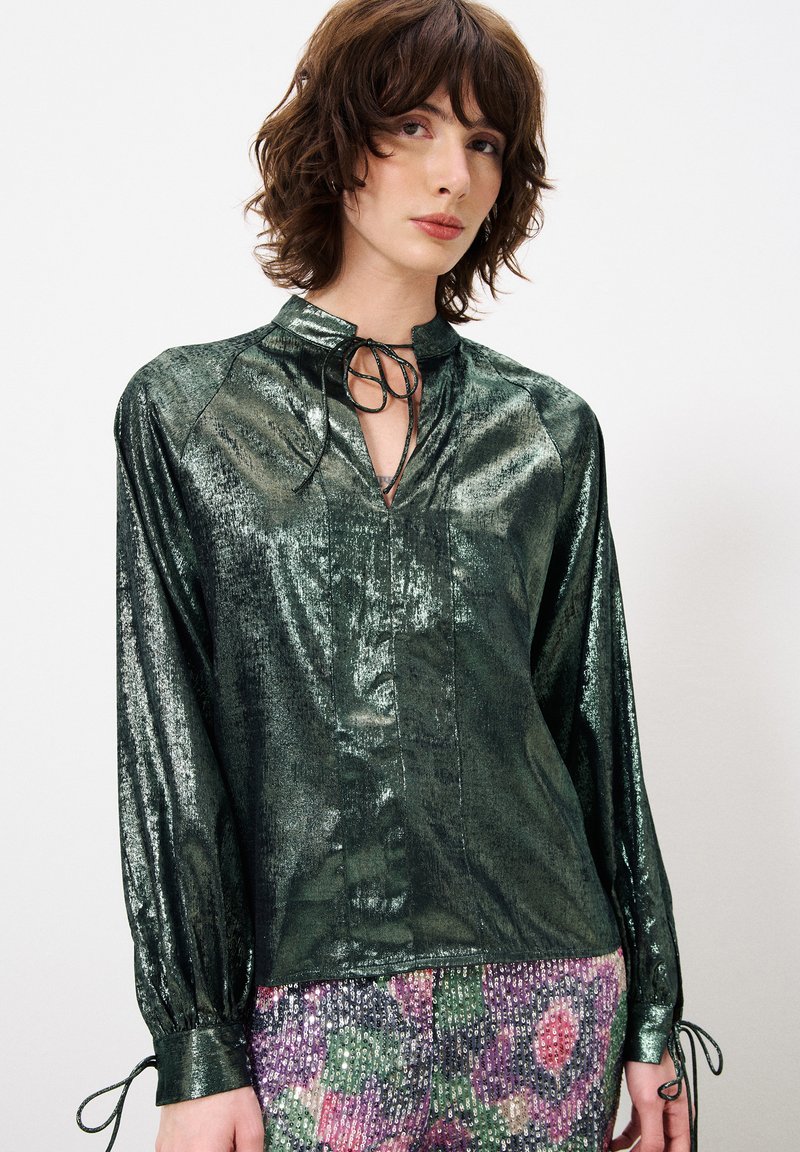 Glanzende groene blouse met een hoge kraag, een sleutelgatopening en een strikdetail. Lange mouwen hebben gathered manchetten. Glad, reflecterend textuur van de stof.