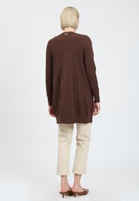 Bruine lange mouwen cardigan met een relaxte pasvorm, gemaakt van zacht materiaal, met een rechte zoom en zijsplitten, gecombineerd met lichtbeige broeken.