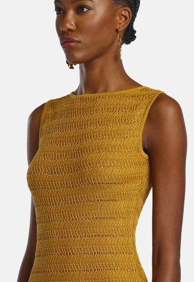 Femme aux cheveux tressés portant des boucles d'oreilles créoles en or et un haut sans manches jaune moutarde en maille texturée, tournée vers la gauche.