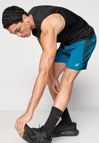 Μαύρο αμάνικο πουκάμισο, αθλητικές βερμούδες τιρκουάζ με πλαϊνή φάσα, μαύρα αθλητικά παπούτσια και μαύρες κάλτσες. Άτομο που διορθώνει το παπούτσι, εστίαση στο πόδι.