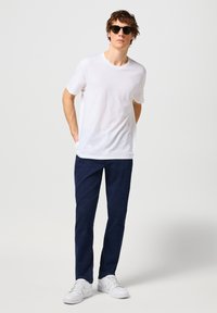 Camiseta blanca de algodón, pantalones de mezclilla azul oscuro y zapatillas blancas. Gafas de sol con lentes oscuros. Ajuste casual con una postura relajada.