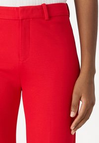 Røde skreddersydde bukser har et glatt, levende stoff, flat front med lommer, og en slank passform akkompagnert av beltehemper.