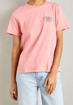 Vaaleanpunainen lyhythihainen puuvillainen t-paita, jossa on pyöreä pääntie ja edessä teksti "CATCH THE WAVE" sekä vaaleansiniset farkut.