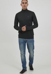Pulover gri cu guler înalt, realizat dintr-un material tricotat, cu un design ajustat și manșete strâmte, asociat cu blugi albaștri deschiși și adidași negri.