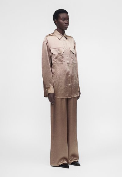 Μοντέλο φοράει φαρδιά, γυαλιστερή μπεζ σατέν πουκάμισο με δύο τσέπες στο στήθος και αντίστοιχο παντελόνι με φαρδιά πόδια, στέκεται με τα χέρια δίπλα στο σώμα.