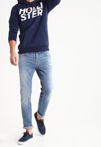 Sudadera navy con gráfico de "HOLISTER", jeans desgastados de color azul claro y zapatillas navy. Presenta un corte relajado y puños enrollados.