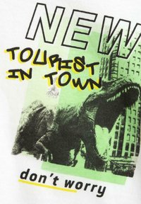 Bílá bavlněná triko s grafickým designem zobrazujícím dinosaura, zelenými akcenty a výrazným textem "NOVÝ TURISTA VE MĚSTĚ" a "nedělej si starosti."