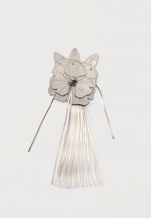 Fleur en cuir argenté avec trois pétales et de longues franges suspendues en dessous sur un fond clair uni.