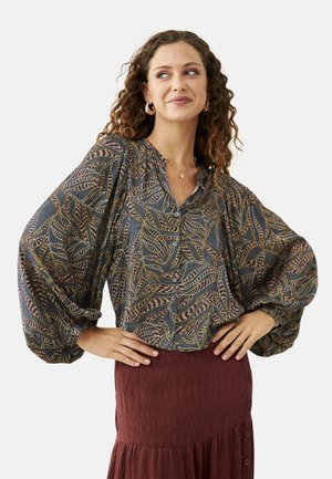 Mujer con cabello rizado que lleva una blusa abotonada con estampado y mangas voluminosas, y una falda texturizada color burdeos con botones laterales.