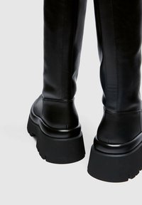 Botas de cuero negras hasta la rodilla con una suela gruesa y texturizada y puntera redondeada. Detalles de costura visibles en la parte superior de la bota.