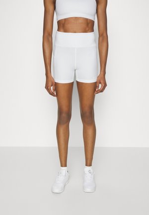 Osoba atletyczna ubrana w białe, wysokie spodenki rowerowe, biały sportowy biustonosz i białe trampki, stojąca na jednolitym tle.