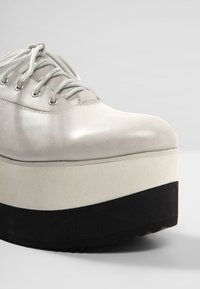 Chaussure plateforme en cuir blanc avec semelle épaisse noire et blanche et lacets blancs sur un fond gris uni.