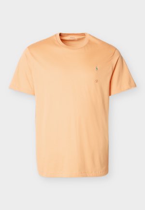 Mann mit Dreadlocks, der ein oranges T-Shirt, hellblaue Jeans und schwarze Turnschuhe trägt, steht mit einer Hand in der Tasche vor einem schlichten Hintergrund.