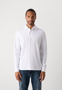 Mann, der ein schlichtes weißes Langarm-Polohemd und eine blaue Jeans trägt, vor einem hellgrauen Hintergrund steht.