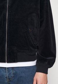 Black corduroy jacket with a zippered front, ribbed cuffs, and a buttoned pocket on the side. Soft texture with vertical stripes.