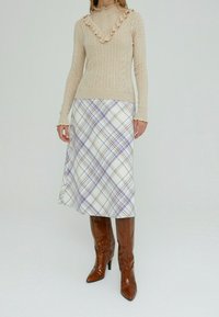 Beige gebreide trui met ruches bij de halslijn en een gestructureerd patroon, gecombineerd met een witte geruite rok met paarse en bruine lijnen. Bruine laarzen tot aan de knie.