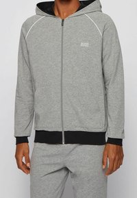 Sudadera gris con cremallera y ribete negro, con una capucha amplia, mangas largas y un logo en el pecho izquierdo. Textura de tela suave.