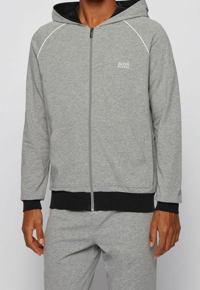 Sudadera gris con cremallera y ribete negro, con una capucha amplia, mangas largas y un logo en el pecho izquierdo. Textura de tela suave.