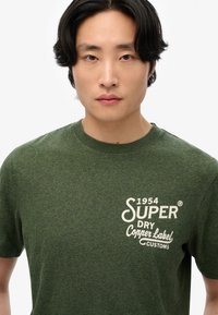 Grøn crew-neck t-shirt lavet af tekstureret stof, med et hvidt logo print, der inkluderer "1954 SUPER DRY Copper Label CUSTOMS."