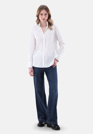 Weißes Hemd mit langem Arm, kombiniert mit dunkelblauer weit geschnittener Jeans. Der Stoff wirkt glatt und leicht. Schwarze Schuhe runden das Outfit ab.