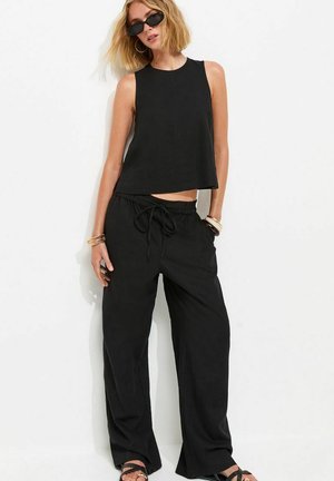 Femme blonde portant un haut noir sans manches, un pantalon noir ample avec cordon de serrage, des lunettes de soleil noires et des bracelets, posant contre un mur blanc.