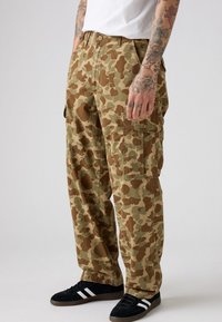 Pantalones cargo con un patrón de camuflaje que presenta colores marrón, verde y marrón claro. Están hechos de una tela con un corte relajado y múltiples bolsillos.
