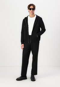 Μαύρη φούτερ ζακέτα με κορδόνια, λευκό μπλουζάκι με στρογγυλή λαιμόκοψη, μαύρο παντελόνι και μαύρα παπούτσια. Λείο ύφασμα, άνετη εφαρμογή, μίνιμαλ σχεδίαση.