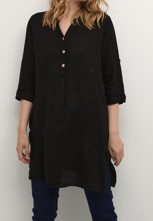 Kvinde iført en løs sort tunika med tre træknapper, opsmøgede ærmer og mørkeblå bukser mod en ensartet hvid baggrund.