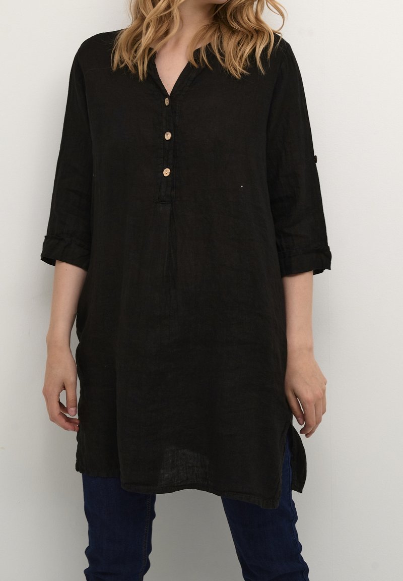 Femme portant une tunique noire ample avec trois boutons en bois, des manches retroussées, et un pantalon bleu foncé sur un fond blanc uni.