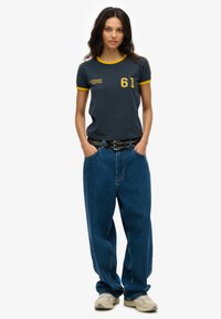 Dark blue t-shirt with yellow trim and logo, paired with loose-fitting blue jeans. Black belt with studs and beige sneakers complete the outfit.