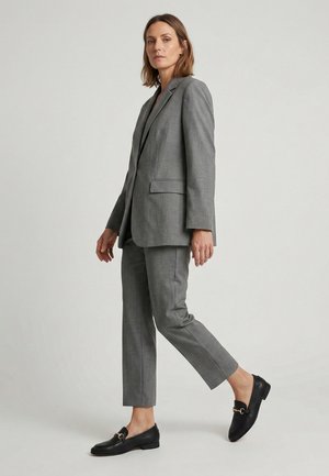 Vrouw loopt, draagt een op maat gemaakte grijze blazer en bijpassende cropped broek, gecombineerd met zwarte leren loafers op een lichte achtergrond.