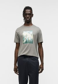 Γκρι βαμβακερό μπλουζάκι με τυπωμένο σχέδιο βουνών και δέντρων. Το μοντέλο φοράει ναυτικό παντελόνι με κορδόνι στη μέση, παρουσιάζοντας άνετη εφαρμογή.