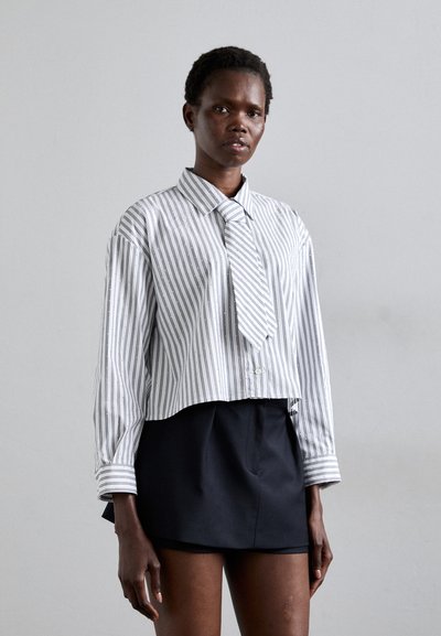 Chemise rayée blanche et bleu clair, à manches longues, avec un col pointu et une cravate assortie, associée à une jupe sombre.