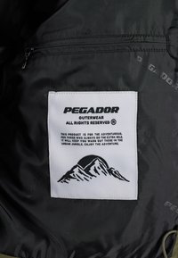 Černá podšívka bundy s bílou štítkem s logem "PEGADOR", grafikou hory a pokyny k údržbě. Obsahuje kapsu na zip.