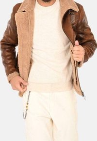 Mężczyzna ubrany w brązową skórzaną kurtkę z kożuchem, na beżowym swetrze i kremowych spodniach z ozdobnym łańcuszkiem na kieszeni.