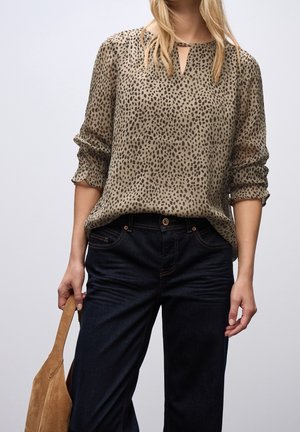 Frau trägt eine beige Leopardenmuster-Bluse mit langen Ärmeln und schwarze Jeans, hält eine hellbraune Wildleder-Handtasche an ihrer Seite.