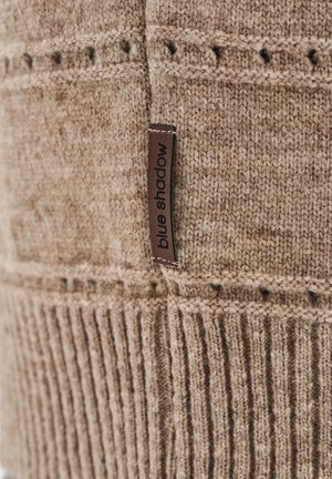 Beige gebreide stof met ribbel- en patroontexturen, met een bruine label genaamd "blue shadow" op het materiaal gestikt.