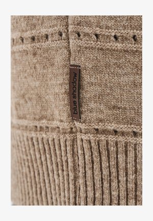 Beżowa dzianina z żeberkowymi i wzorzystymi teksturami, z brązową metką z napisem "niebieski cień" przyszytą do materiału.