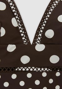 Tissu marron avec de grands pois blancs, doté d'un décolleté en V profond et d'une bordure en dentelle marron décorative le long du col et de la taille.