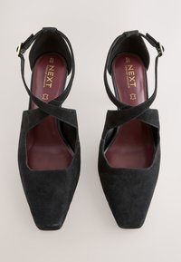 Czarne zamszowe buty na wysokim obcasie z kwadratowym noskiem, z krzyżującymi się paskami oraz smukłym paskiem wokół kostki z złotą klamrą.