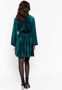 Teal sammetklänning med långa ärmar, samlad midja och utsvängd kjol; använd med svarta strumpbyxor och klackade ankelstövlar.