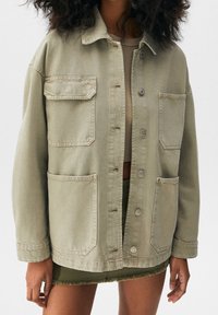 Mujer con una chaqueta beige oversized con cuatro bolsillos frontales sobre un top corto y una falda mini verde con dobladillo deshilachado.