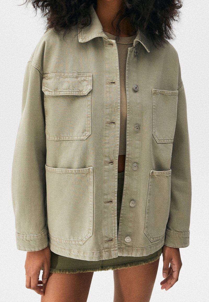 Mujer con una chaqueta beige oversized con cuatro bolsillos frontales sobre un top corto y una falda mini verde con dobladillo deshilachado.