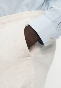 Ανοιχτό μπεζ παντελόνι λινό με ελαστική μέση, που διαθέτει πλαϊνή τσέπη. Φαίνεται μερικώς ένα ανοιχτό μπλε μακρυμάνικο πουκάμισο με λεπτομέρεια κουμπιού.