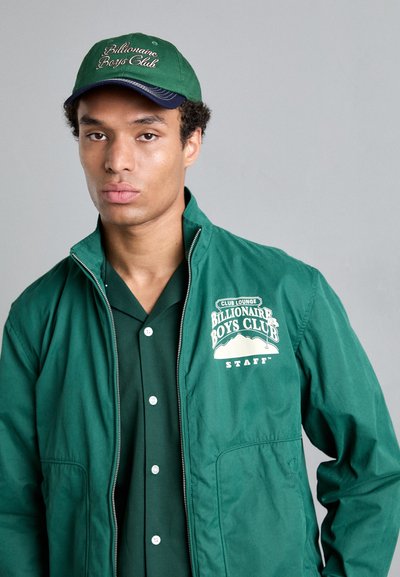 Grüne Jacke mit einem Frontreißverschluss, mit dem Logo "Billionaire Boys Club". Kombiniert mit einem grünen Shirt und einer grünen Baseballmütze mit navyfarbenen Akzenten.