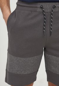 Los pantalones cortos grises están confeccionados con una mezcla de paneles de tela suave y texturada. Cuentan con una cintura elástica con cordones ajustables y bolsillos frontales.