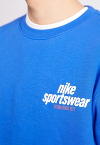 Blå sweatshirt med rund halsringning, tillverkad av mjukt tyg. Har vit text "nike sportswear" och "ETABLERAD 1972" i orange.