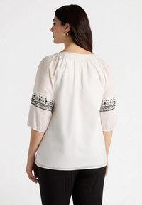 White blouse with three-quarter length puff sleeves, featuring black embroidered band around the sleeves. Smooth texture, relaxed fit.