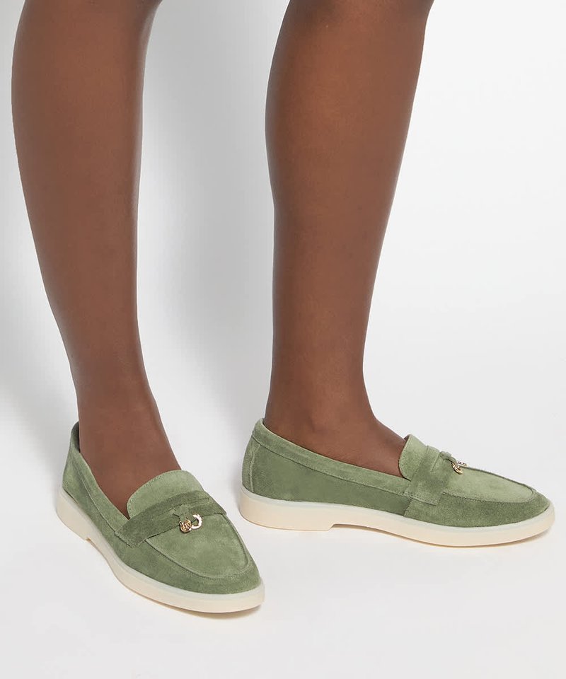 Paire de mocassins verts en daim à enfiler avec semelles crème et petit détail de boucle dorée, portés sur des jambes brun moyen.